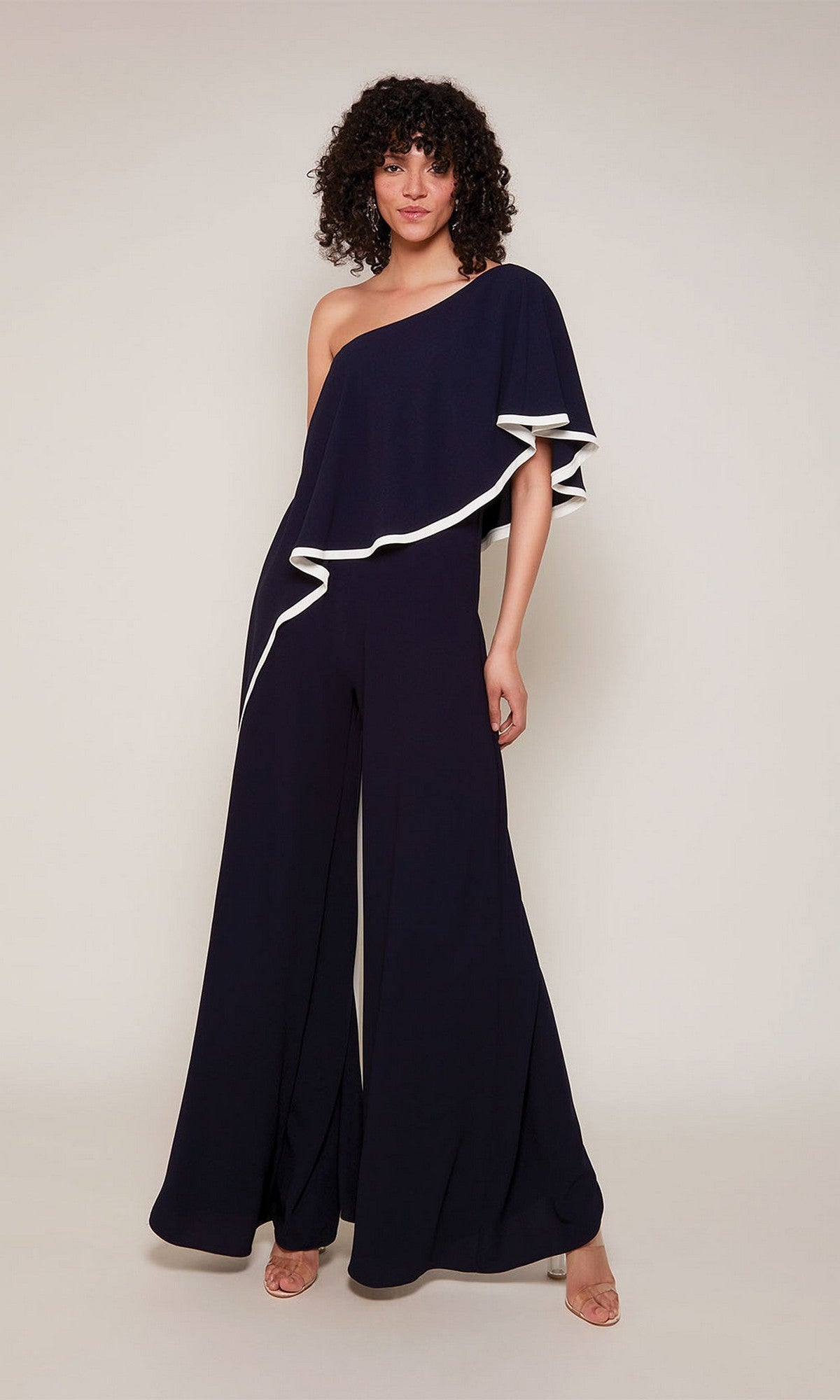 A woman poses in the Alyce 70049 long formal jumpsuit, featuring a midnight blue one-shoulder design with diamond white trim and wide legs, styled with nude heels against a plain light background.